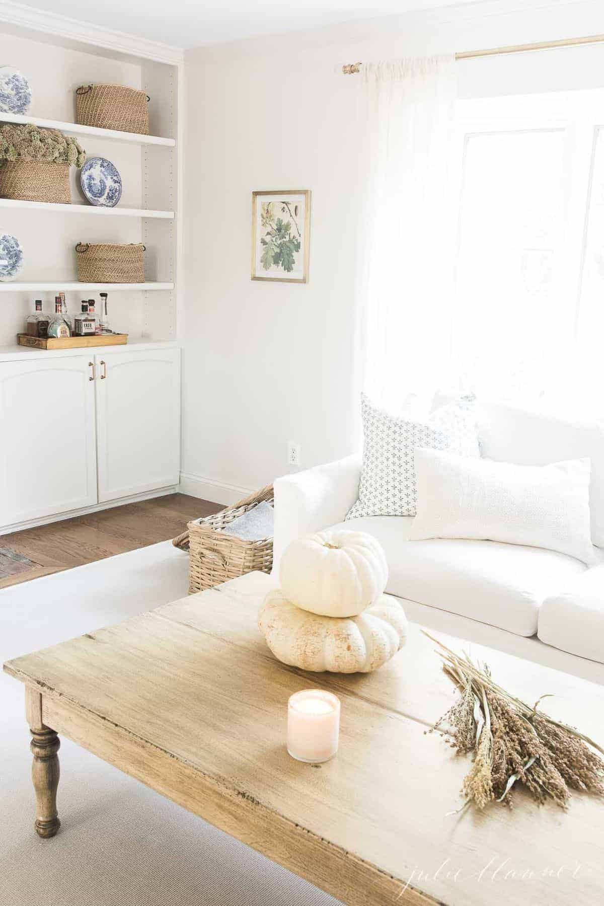 客厅设有白色沙发，建立在书架，和秋天的装饰，如蜡烛，秋天的树叶和白色南瓜在前景的咖啡桌上。# fallliviingroom # harvestdecor
