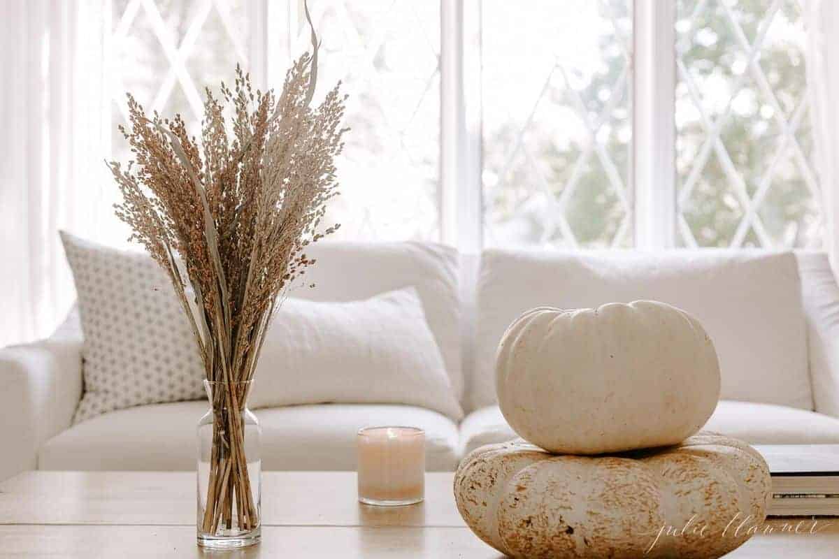 木制的咖啡桌，白色的沙发和窗户为背景，白色的南瓜，蜡烛和花瓶秋天的树叶作为装饰。# falllivingroom # harvestdecor