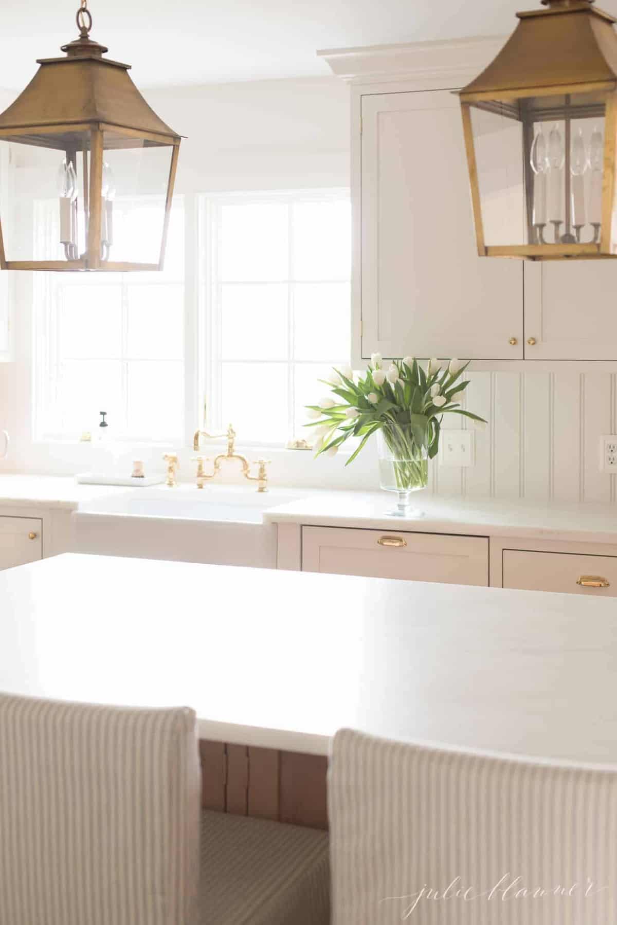 一个白色的厨房，带有大窗户，可为房间增添光，岛上的黄铜灯笼。